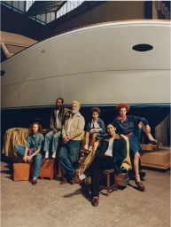 Group of six diverse models wearing casual denim and boat shoes posing in front of a large white yacht inside a warehouse
