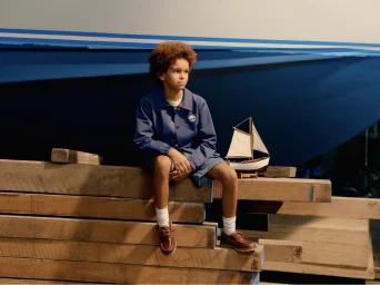 Child seated on stacked wooden beams wearing a blue jacket and boat shoes next to a model sailboat.