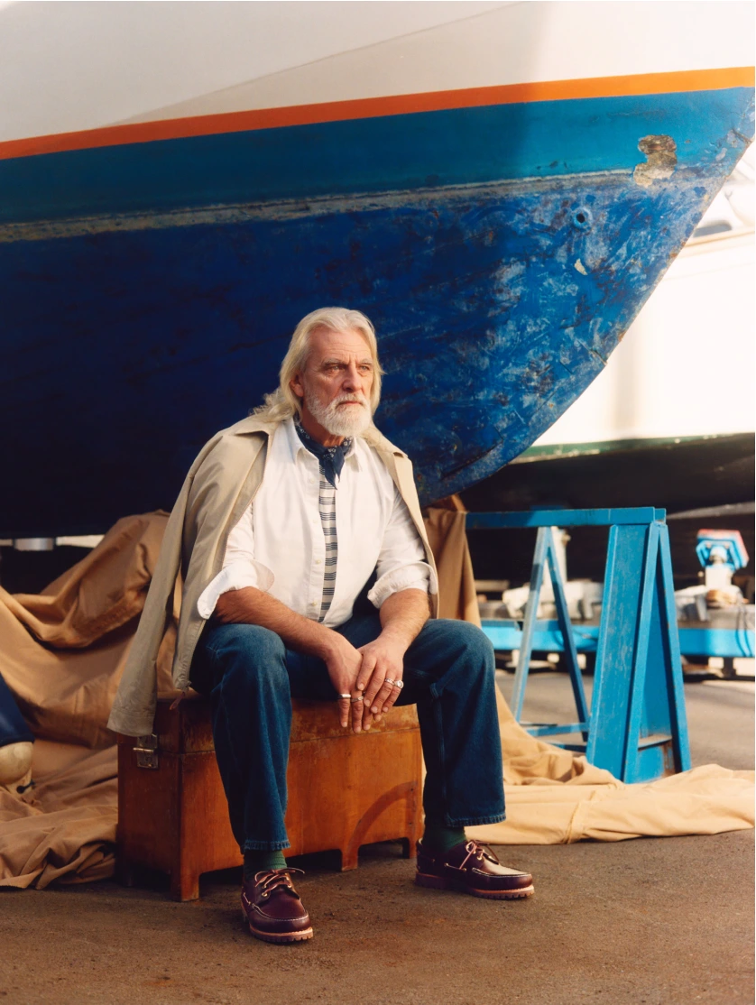 Elderly man with white hair and beard seated on a wooden crate in front of a blue boat hull wearing boat shoes.