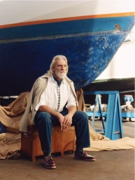 Elderly man with white hair and beard seated on a wooden crate in front of a blue boat hull wearing boat shoes.