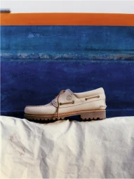 Cream leather boat shoe rests on a textured white fabric surface against a blue and orange backdrop.