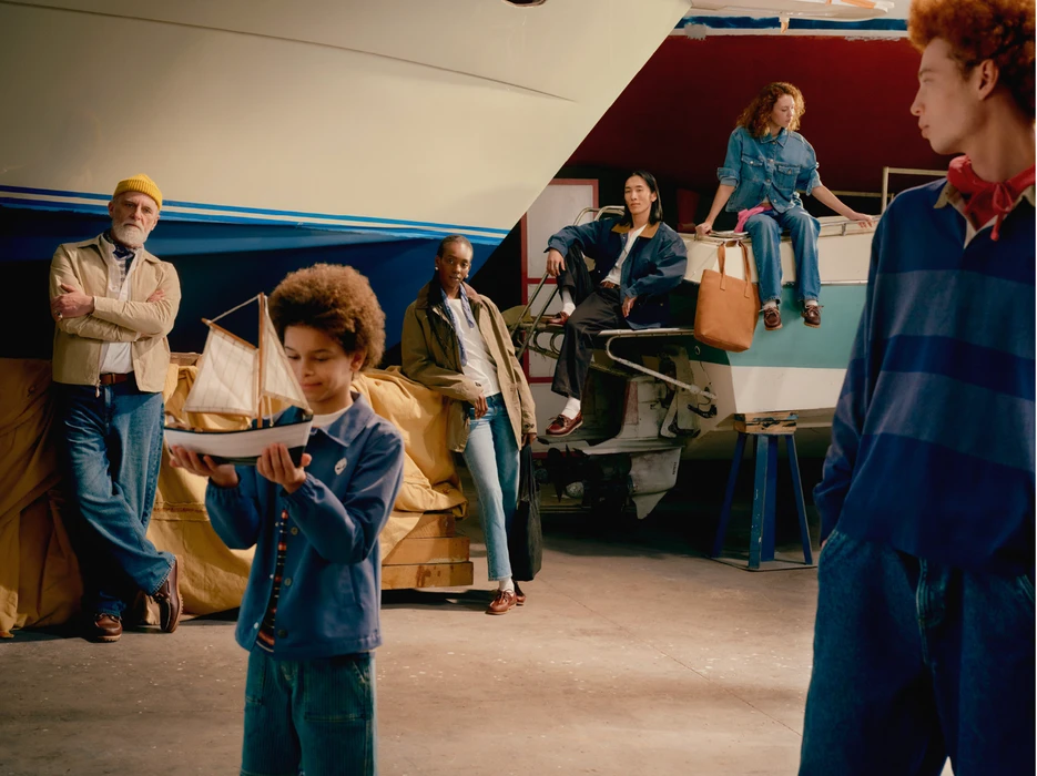 Group of models posing in an indoor boathouse or garage with a boat hull, holding a model sailboat.