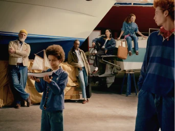 Group of models posing in an indoor boathouse or garage with a boat hull, holding a model sailboat.