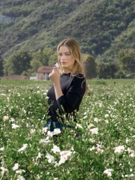Margot Robbie holding a small white flower in a lush green floral field with mountains in the background
