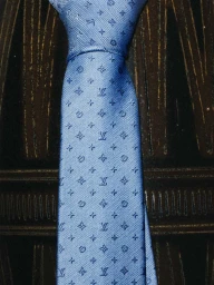 Close-up of a blue silk necktie with a repeating monogram pattern against a dark textured background.