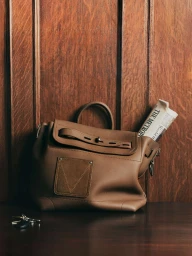 Brown leather handbag resting on a dark wooden surface in front of wood paneling with keys and a newspaper tucked inside
