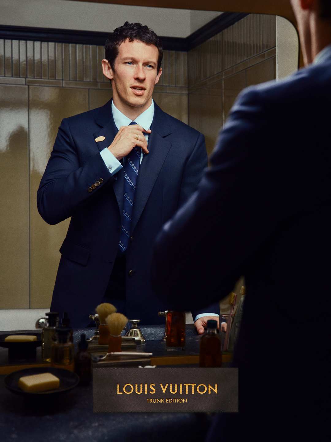 Man adjusting blue tie in a bathroom mirror wearing a navy suit jacket and light blue collared shirt.