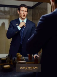 Man adjusting blue tie in a bathroom mirror wearing a navy suit jacket and light blue collared shirt.