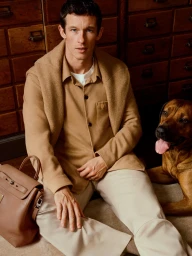 A man in a tan shirt and sweater sitting with a brown dog and a leather backpack indoors against wooden drawers.