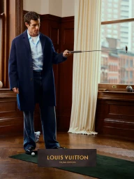Man wearing a navy coat holding a golf putter indoors on a putting mat with wood-paneled walls.