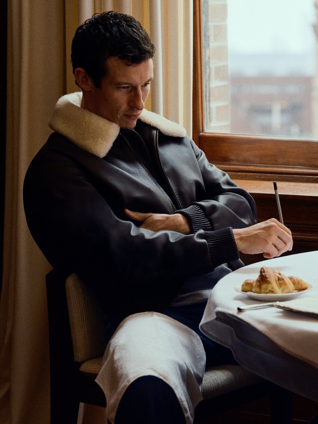 Man wearing a dark leather jacket with a white shearling collar seated at a table indoors near a window.