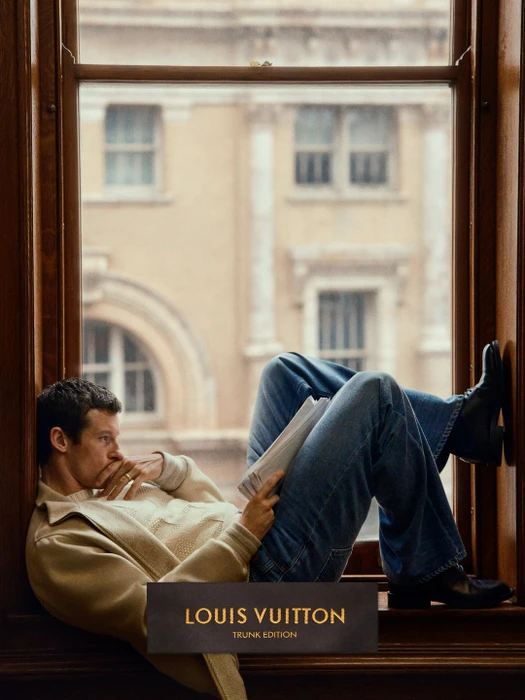 Man in cream sweater and blue jeans sitting on a large wooden windowsill reading papers