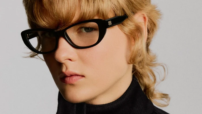 Close-up portrait of a model wearing thick black cat-eye glasses and a black turtleneck against a plain light background