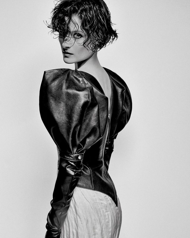 Monochrome fashion shot of a woman with a wet curly hairstyle in a leather top with puffy shoulders and long sleeves, posing in a photo studio.