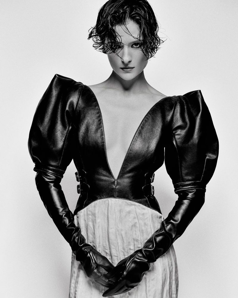 Monochrome fashion portrait of a woman with short hair, wearing a black leather top with large sleeves and long gloves against white background.