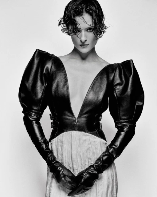 Monochrome fashion portrait of a woman with short hair, wearing a black leather top with large sleeves and long gloves against white background.