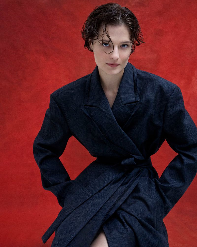 Woman with short dark hair poses in a wrap-style blue blazer against a textured red backdrop in a studio portrait.