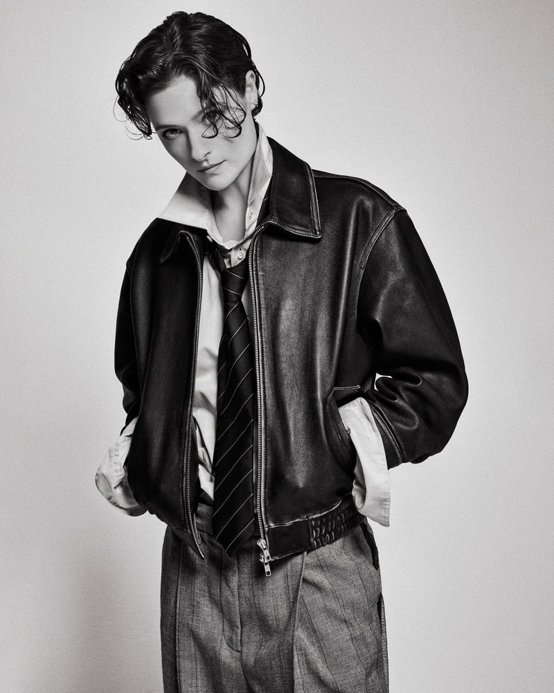 Monochrome studio portrait of a young adult wearing a leather jacket, collared shirt, and tie, hands in pockets, against a neutral background.