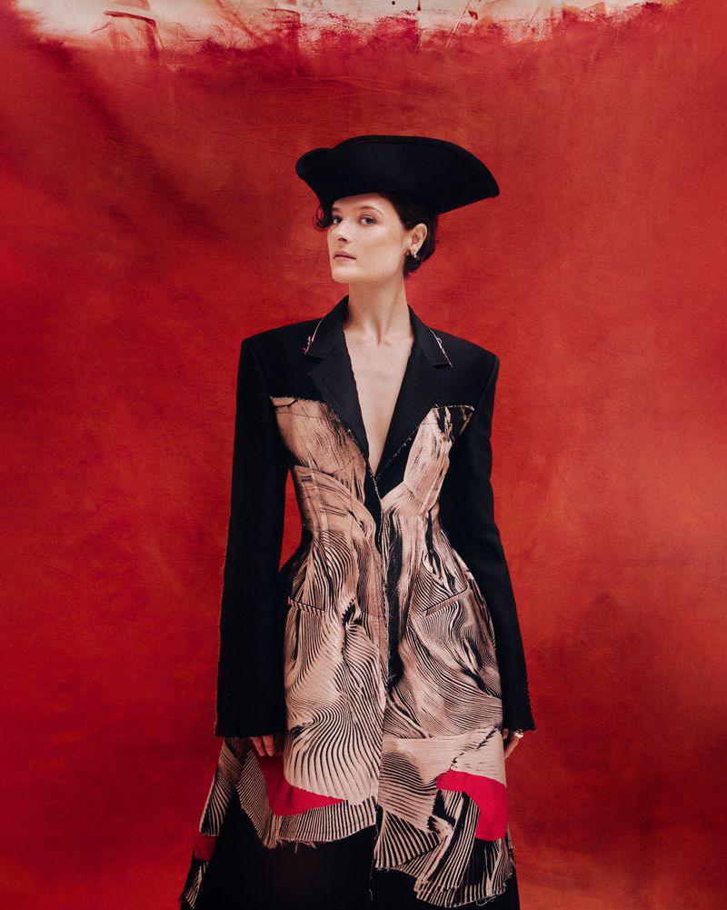 A woman in a black sculptural hat and a black jacket with an abstract beige print poses against a mottled red studio backdrop.