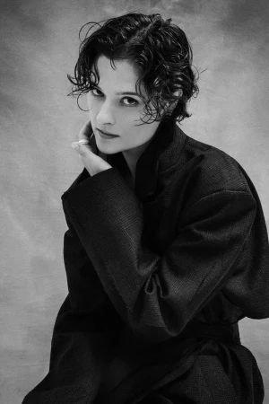 Black and white portrait of a person with short curly hair wearing an oversized dark coat against a mottled backdrop.