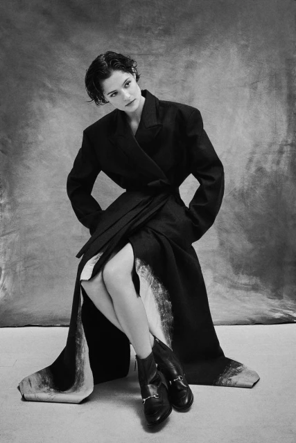 Monochrome portrait of an adult female in a dark wraparound coat and boots, seated against a mottled gray background in a studio.