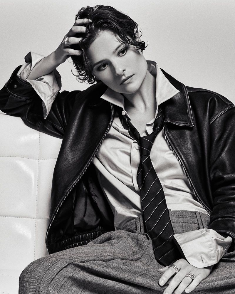 Black and white portrait of adult female in a leather jacket, collared shirt, striped tie, and grey slacks leaning on white cushion.