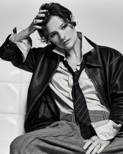 Black and white portrait of adult female in a leather jacket, collared shirt, striped tie, and grey slacks leaning on white cushion.