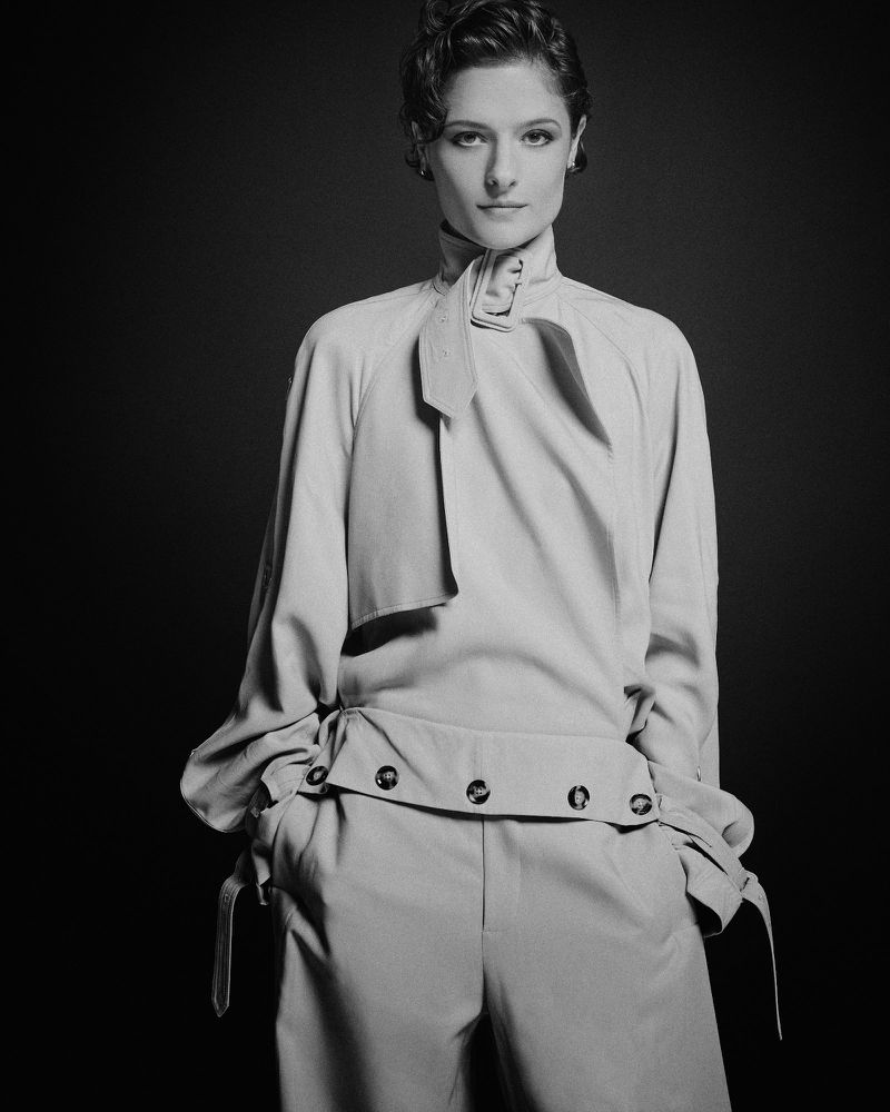 Monochrome fashion portrait of a woman in a light-colored, long-sleeved blouse and trousers, posing against a dark backdrop in a studio.