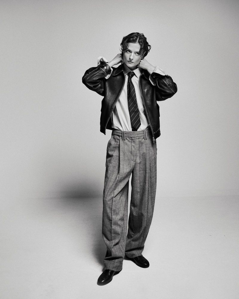Black and white portrait of a model wearing a leather jacket, white shirt, tie, and wide-legged trousers in a studio setting.