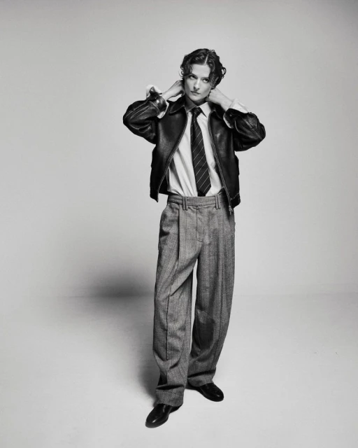 Black and white portrait of a model wearing a leather jacket, white shirt, tie, and wide-legged trousers in a studio setting.