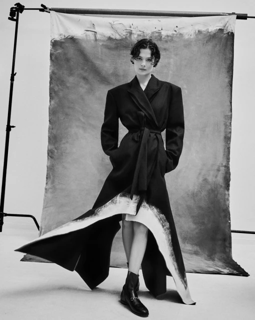 Elegant woman in a black wrap coat poses in a photo studio with a gray painted canvas backdrop; she is wearing black ankle boots.