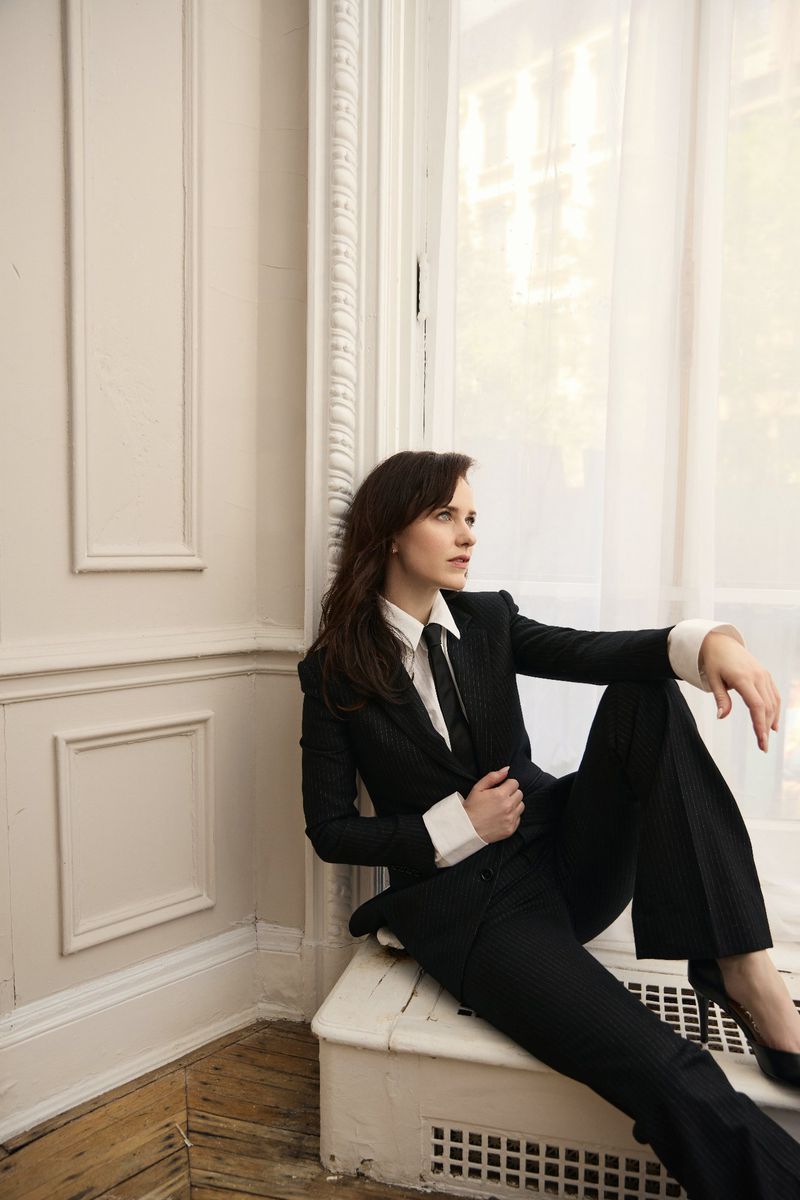 Elegant woman in a black pantsuit poses near a window with natural light. She is seated and gazing off-camera, with a calm facial expression.