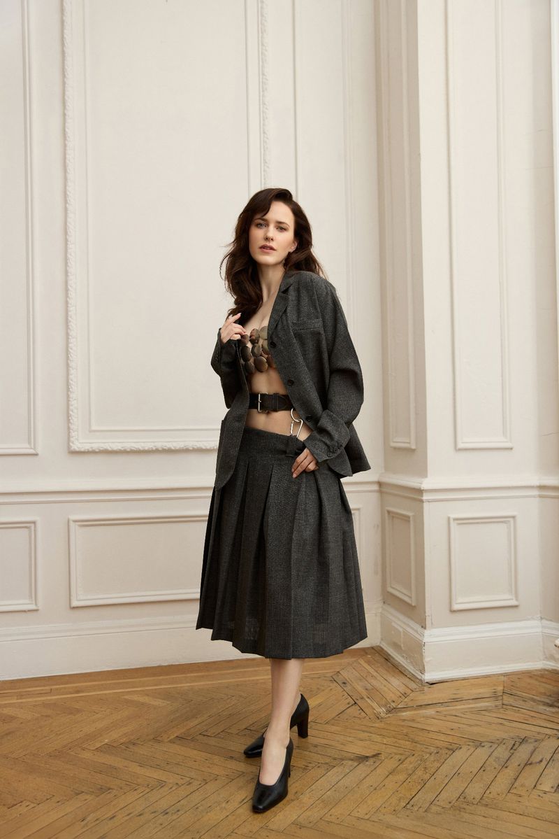 Brunette woman posing indoors wearing a grey skirt suit, bra top, and black high heels in a fashion-editorial style full-length studio shot.
