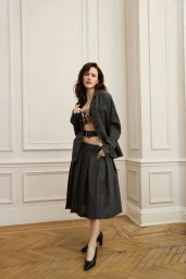 Rachel Brosnahan in a dark grey suit jacket and pleated midi skirt standing against a white paneled wall on herringbone wood flooring.