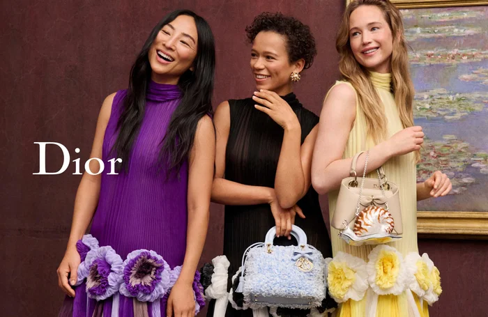 Three female models wearing vibrant dresses against a textured dark wall holding designer handbags for Dior Cruise 2027