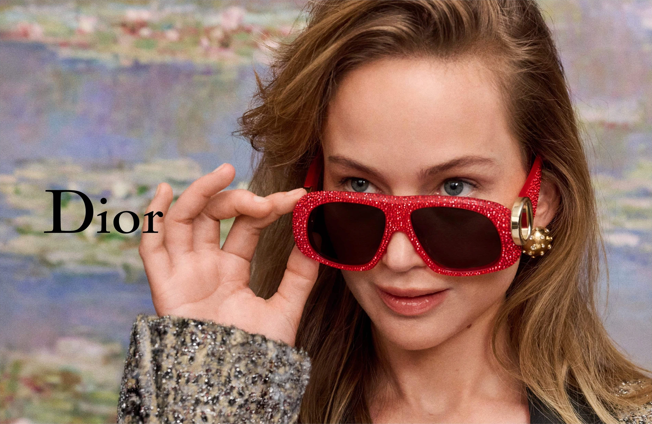 Model wearing red crystal sunglasses adjusting frames against an impressionistic water-lily backdrop