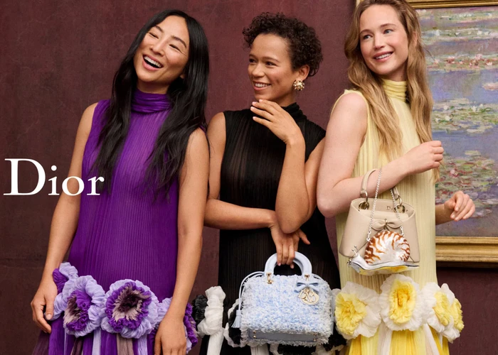 Three models wearing colorful pleated dresses and holding handbags in front of a dark burgundy wall with framed art.