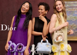 Three models wearing colorful pleated dresses and holding handbags in front of a dark burgundy wall with framed art.