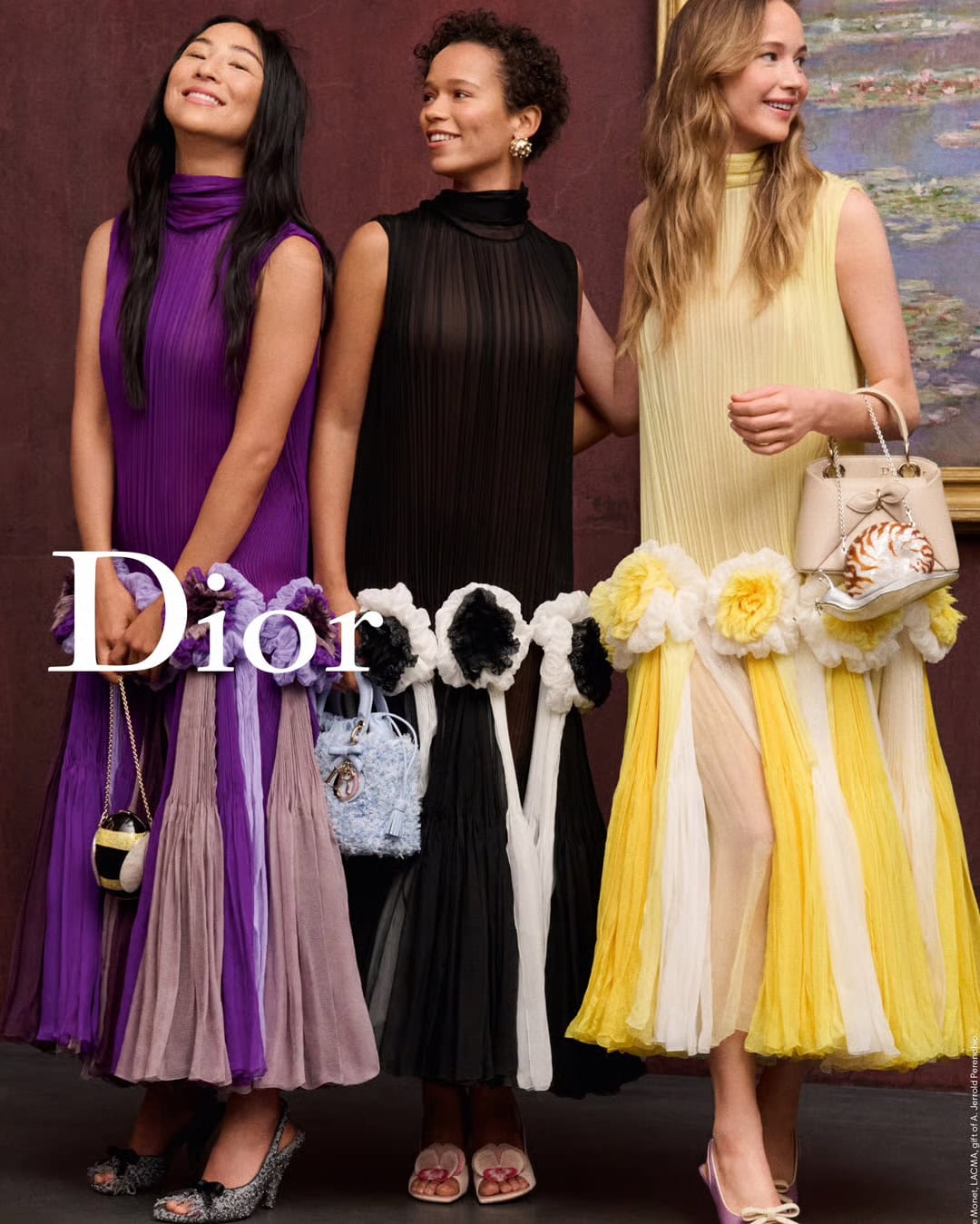 Three models wearing colorful pleated dresses with floral appliques and holding handbags in an indoor setting