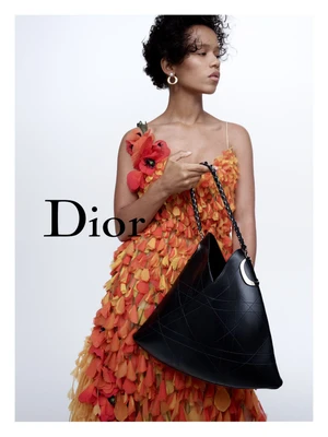 Model in textured orange petal dress holding a black leather handbag against a plain white background with Dior logo