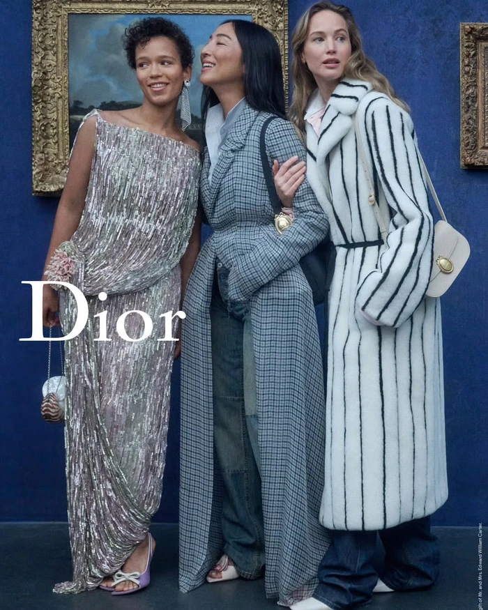 Three models in designer apparel pose against a deep blue wall with framed artwork for the Dior Cruise 2027 campaign.