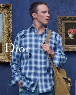 Drew Starkey wearing a blue plaid shirt with numbers and a tan bag against a dark blue textured wall with framed paintings