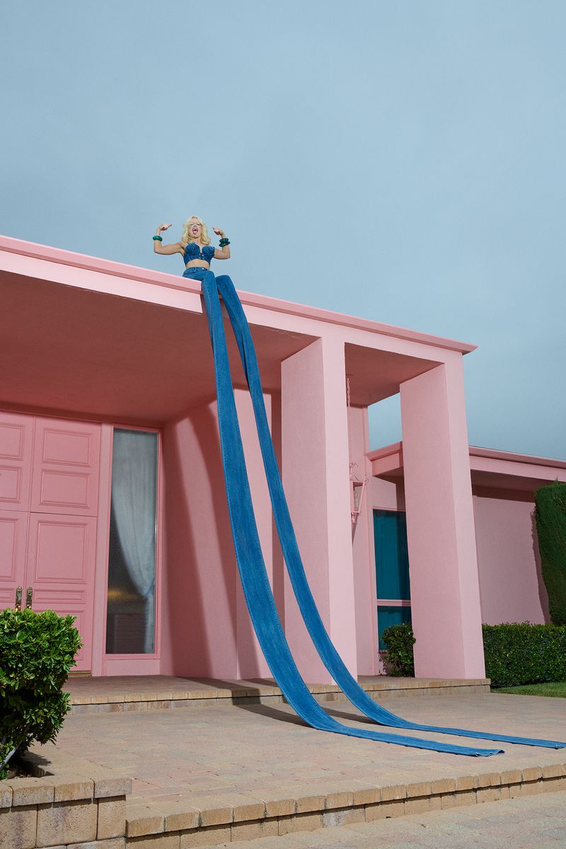 A woman sits on the roof of a pink house, wearing a blue top and long blue fabric extensions in an outdoor setting on a sunny day.