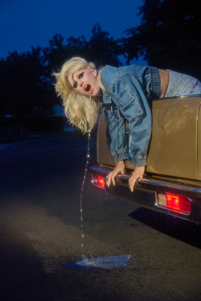 Blonde woman in denim jacket leaning out of a gold car trunk with water dripping from her mouth, parked on a street at dusk.