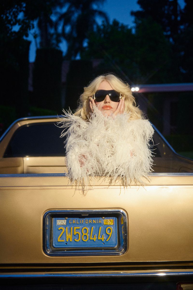 Blonde woman in feathered coat poses in gold vintage car at dusk. Sunglasses and palm trees create a California mood.
