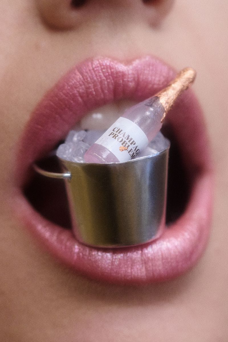 Close-up of a person's mouth holding a miniature champagne bottle in an ice bucket, set between the lips, emphasizing beauty and indulgence.