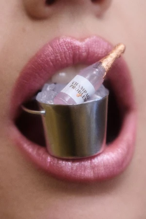 Close-up of a person's mouth holding a miniature champagne bottle in an ice bucket, set between the lips, emphasizing beauty and indulgence.