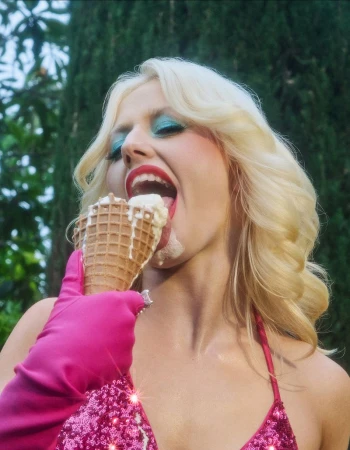 A woman with platinum blonde hair in a pink sequined bikini eating an ice cream cone outdoors with green foliage in the background.