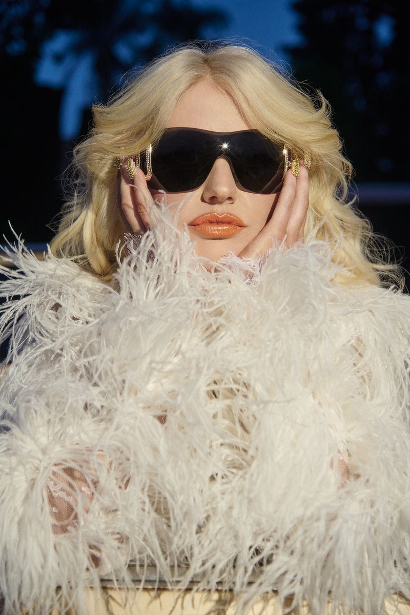 Stylish woman with blonde wavy hair poses with oversized sunglasses, hands to face, wearing feathered boa in a dark outdoor setting.
