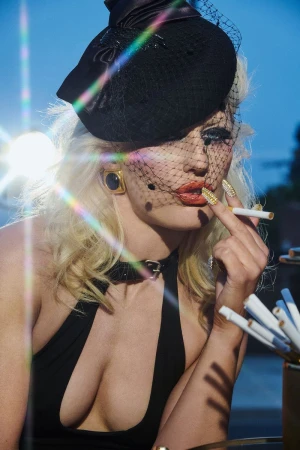 A woman with platinum blonde hair and a black hat is smoking a cigarette with a collection of cigarettes in the foreground.